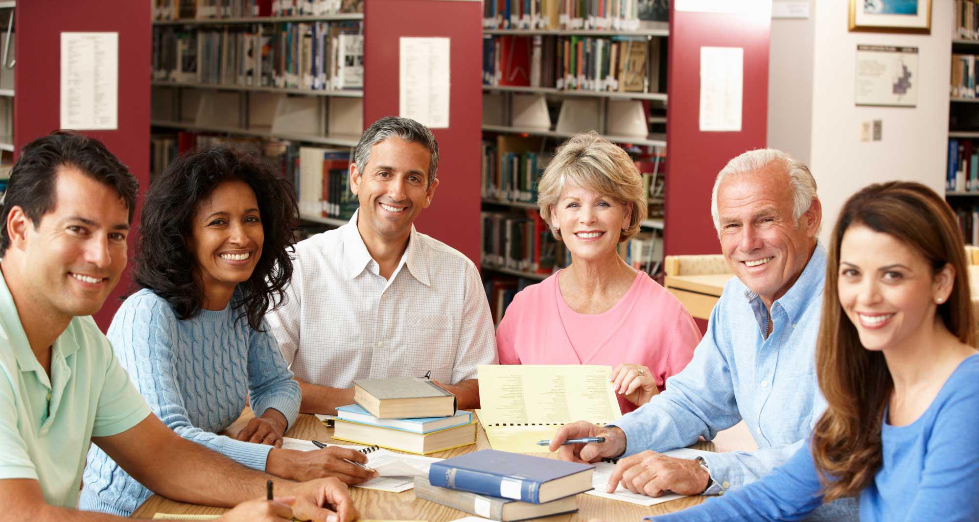 A diverse group of people sitting at a library table, smiling and looking at the camera, with books and notebooks spread out in front of them.